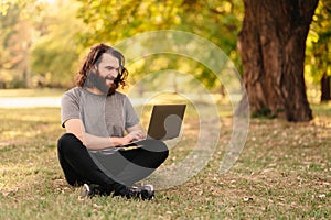 Handsome bearded man with long hair is using his laptop while sitting on grass.