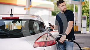 Handsome bearded man refueling car and looking on the scoreboard while standing on self service gas station