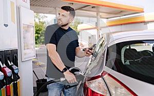 Handsome bearded man refueling car and looking on the scoreboard while standing on self service gas station