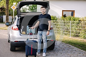 Handsome bearded man stand with suitcase in front of open car trunk. Travel road trip. Holiday summer vacation concept