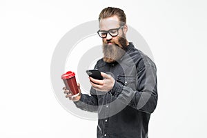Handsome bearded man with stylish hair beard and mustache use mobile phone holding cup or mug drinking tea or coffee on white