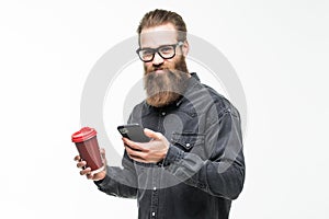 Handsome bearded man with stylish hair beard and mustache use mobile phone holding cup or mug drinking tea or coffee on white