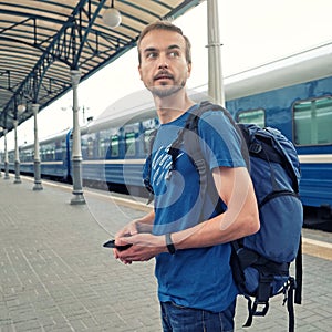 Handsome bearded man tourist with backpack stand on railway station platform and waiting for train. Travel concept. Square