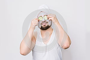 Handsome bearded man with towel on head and whilte clay mask on skin, covers his eyes with two slices of cucumber. Spa