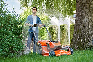 Handsome bearded man trimming lawn with electric mower, working on backyard at sunny day.
