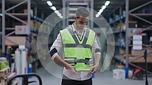 Handsome bearded Middle Eastern man tying velcro on vest standing in industrial warehouse and looking around. Portrait