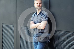 Handsome bearded millennial casual wearing man with stylish leather bag standing near black wall in the city streeet photo