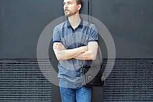 Handsome bearded millennial man in polo shirt and jeans with stylish leather bag standing near black wall in the city streeet photo