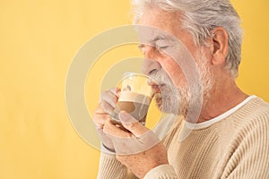 Handsome bearded senior man drinking coffee and milk cappuccino from a glass beaker , standing on yellow background