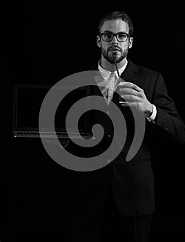 Handsome bearded stylish business man in elegant black suit in glasses holding laptop and coffee cup in studio on black