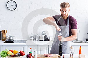 handsome bearded young man in apron opening bottle of wine and looking at camera while cooking