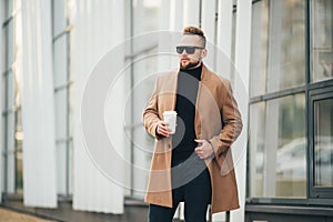 Handsome bearded young man in sunglasses holding cup of takeaway coffee near business center.