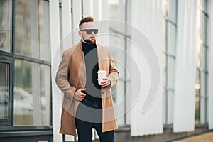 Handsome bearded young man in sunglasses holding cup of takeaway coffee near business center.