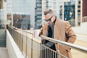 Handsome bearded young man in sunglasses holding cup of takeaway coffee near business center.