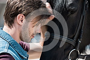 Handsome bearded young man touching purebred brown horse outdoors