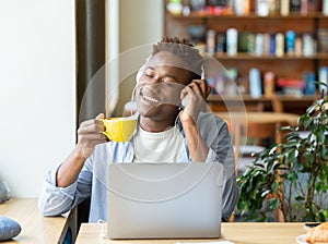 Handsome black guy with headphones listening to music with closed eyes and drinking coffee at cozy cafe