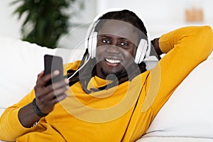 Handsome Black Guy With Smartphone And Headphones Relaxing On Couch At Home