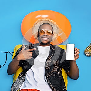 Handsome black guy in summer wear sunbathing on inflatable lilo, pointing at smartphone with white empty screen, mockup