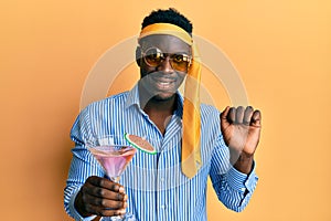 Handsome black man drunk wearing tie over head and sunglasses drinking a cocktail screaming proud, celebrating victory and success