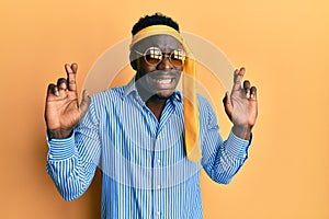 Handsome black man drunk wearing tie over head and sunglasses gesturing finger crossed smiling with hope and eyes closed