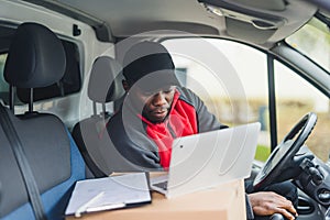 Handsome Black man in his 30s sitting inside a delivery van and checking parcel documents on silver laptop. Courier