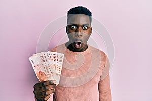 Handsome black man holding 100 new zealand dollars banknote scared and amazed with open mouth for surprise, disbelief face
