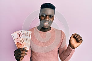 Handsome black man holding 100 new zealand dollars banknote screaming proud, celebrating victory and success very excited with