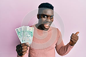 Handsome black man holding 100 polish zloty banknotes pointing thumb up to the side smiling happy with open mouth