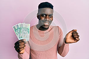 Handsome black man holding 100 polish zloty banknotes screaming proud, celebrating victory and success very excited with raised