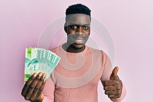 Handsome black man holding 20000 indonesian rupiah smiling happy and positive, thumb up doing excellent and approval sign