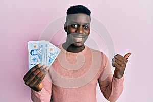 Handsome black man holding 50 thai baht banknotes pointing thumb up to the side smiling happy with open mouth