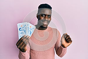 Handsome black man holding 50 thai baht banknotes screaming proud, celebrating victory and success very excited with raised arm