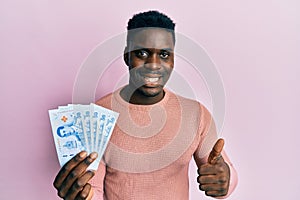 Handsome black man holding 50 thai baht banknotes smiling happy and positive, thumb up doing excellent and approval sign