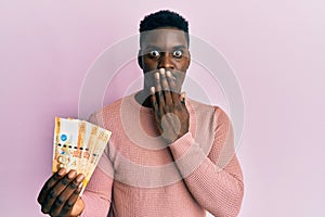 Handsome black man holding 500 philippine peso banknotes covering mouth with hand, shocked and afraid for mistake