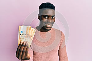 Handsome black man holding 500 philippine peso banknotes looking positive and happy standing and smiling with a confident smile