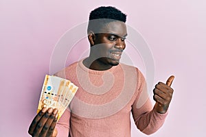 Handsome black man holding 500 philippine peso banknotes pointing thumb up to the side smiling happy with open mouth