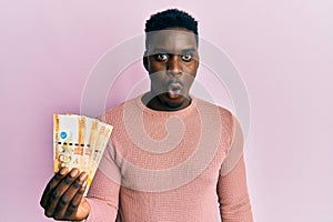 Handsome black man holding 500 philippine peso banknotes scared and amazed with open mouth for surprise, disbelief face