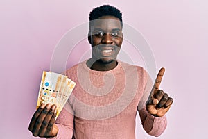 Handsome black man holding 500 philippine peso banknotes smiling with an idea or question pointing finger with happy face, number