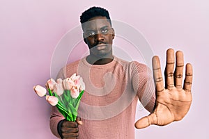 Handsome black man holding bouquet of pink tulips flowers with open hand doing stop sign with serious and confident expression,