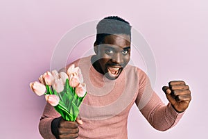 Handsome black man holding bouquet of pink tulips flowers screaming proud, celebrating victory and success very excited with