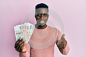 Handsome black man holding czech koruna banknotes smiling happy and positive, thumb up doing excellent and approval sign