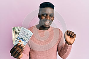 Handsome black man holding dollars screaming proud, celebrating victory and success very excited with raised arm