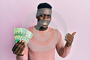 Handsome black man holding south african 10 rand banknotes pointing thumb up to the side smiling happy with open mouth