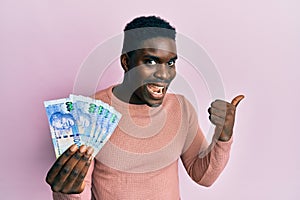 Handsome black man holding south african 100 rand banknotes pointing thumb up to the side smiling happy with open mouth