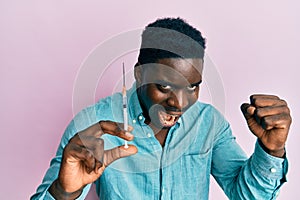 Handsome black man holding syringe screaming proud, celebrating victory and success very excited with raised arms