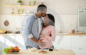 Handsome black man hugging his beautiful pregnant wife and kissing her on forehead in kitchen