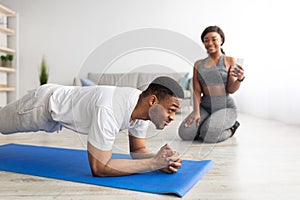 Handsome black man standing in elbow plank on sports mat, doing abs exercise, his girlfriend timing him on cellphone