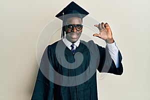 Handsome black man wearing graduation cap and ceremony robe smiling and confident gesturing with hand doing small size sign with