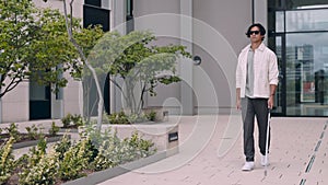 Handsome blind man with disability walking alone on city street with white cane in hands. Concept of special people and