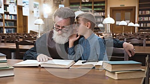 Handsome blond boy whispering to his experienced old grandfather which sitting in the library